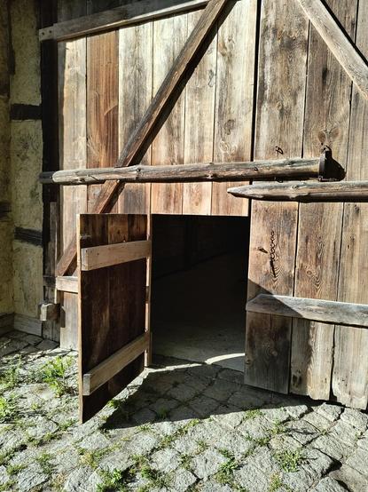 Eine Holzwand aus Brettern, die links an eine Wand aus Fachwerk angrenzt. Im unteren Teil steht eine kleine, halbhohe Holztür offen. Davor liegt das Kopfsteinpflaster mit grün bewachsenen Fugen in der Sonne.