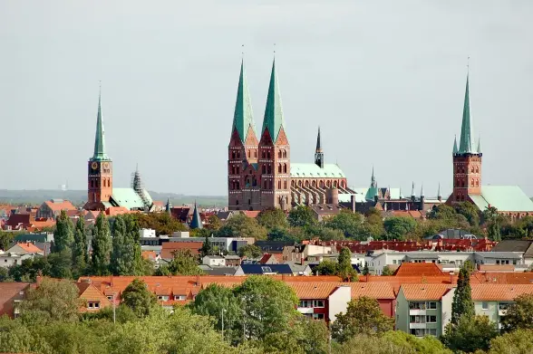 The image depicts a cityscape featuring several towers with pointed roofs and a mix of architectural styles. The prominent buildings include structures with green spires, likely representing churches or historical landmarks. In the foreground, there are residential areas with houses that have orange rooftops, along with trees and vegetation. The overall setting appears to be a European city, characterized by its historical architecture and urban landscape.

Image Credits: Wikimedia / Oleg Dejan / CC BY 3.0