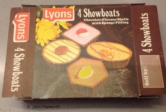 Photograph by John Papworth of an empty cake box from the 1970s. The brown box has a colour picture on the front of four different cupcake-style confections—one of them square, one oval, one round, and one hexagonal—each a different colour and decorated with stripes, swirls, a chocolate drop, jelly sweet etc. Logo of the Lyons brand, white text on red background. Text: 4 Showboats—Chocolate Flavour Shells with Sponge Filling.