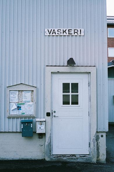 A bleak colour photograph of the door to what might look like an annex to an apartment building from the 50s or 60s. The worn concrete building has grey and wavy metal cladding and an old, faded sign saying ‘vaskeri’, which looks like it might have been hand-painted. The white door itself looks newer, with a small window to the inside. Next to the door is a small billboard for advertisements and notifications and two padlocked postboxes.