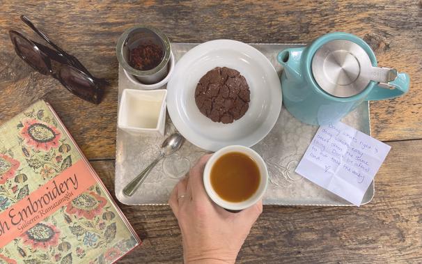 Photo of a tray with a blue teapot, strainer, creamer, white plate with a brown cookie, and a hand holding a cup of tea. To the left of the tray is a pair of black sunglasses and part of a book cover for a book on Turkish Embroidery. There is also a handwritten note on the tray that reads "Doing whats right &amp; doing what's easy aren't often the same thing. Don't be afraid to not take the easy road."