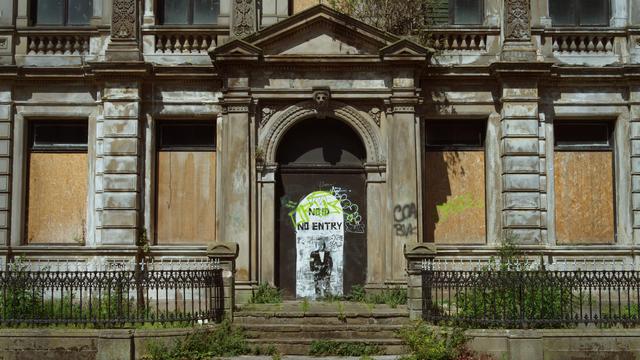 Disused stone clad building on Becket Street, Derby, the windows have been boarded up and the boarded up entrance has some street art of a white archway with a suited person with the words No ID, No Entry above them.