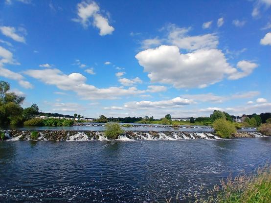 Zwei recht flache Wehre aus Steinen in einem hier breiten Fluss. Blauer Himmel mit weißen Wolken.