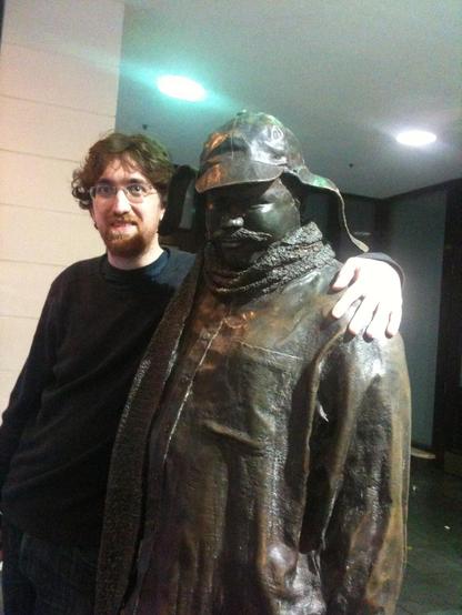 A much younger me stands with his arm around a life-sized statue of a rotund man with a mustache who is wearing a hat with earflaps, a scarf, and a coat. It is Ignatius J. Reilly.