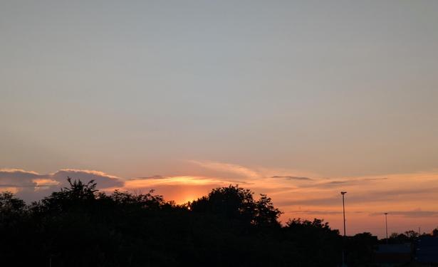 cielo dominato da tonalità calde e sfumate. L'orizzonte è tinto di arancione intenso, indicando la posizione del sole calante, mentre salendo verso l'alto i colori si dissolvono in un rosa tenue e infine in un azzurro-grigio pallido. La silhouette scura di alberi e vegetazione in primo piano crea un netto contrasto con i colori brillanti del cielo, e si intravedono anche alcuni lampioni,