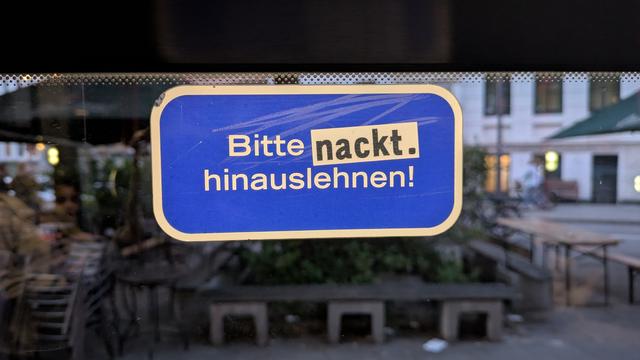 Blauer Aufkleber mit weißem Rand am Busfenster eines Busses des öffentlichen Nahverkehrs einer Großstadt. 
Aufschrift:
Bitte nackt. hinauslehnen!
Der Aufkleber liest sich eigentlich:
Bitte nicht hinauslehnen!