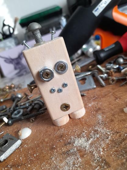 Dřevěná dětská kostka/hranolek jako postavička, robotek. Oči, pusa, ručičky a anténky jsou ze šroubků, nožičky tvoří dřevěná kolečka.