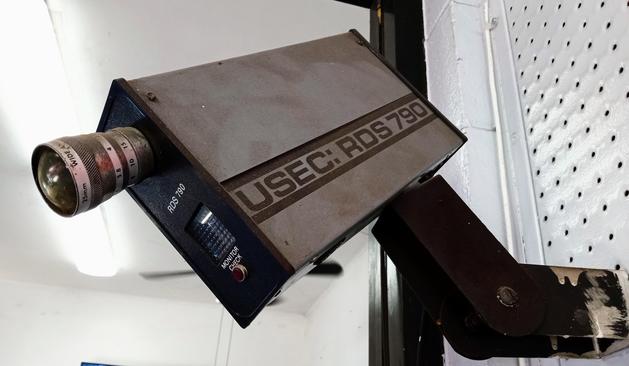 A very boxy and dusty 70s or 80s era security camera, mounted on an angular arm high up on a white office wall, obviously no longer working. It has a large protruding lens, and on the side in big block letters it's labeled "USEC: RDS 790"