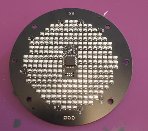 Black circular circuit board approximately 9cm in diameter, with a grid of over 300 LEDs on, also in a circle. There is a rectangle in the middle with no LEDs, like a pupil, but with a microcontroller and other small components.