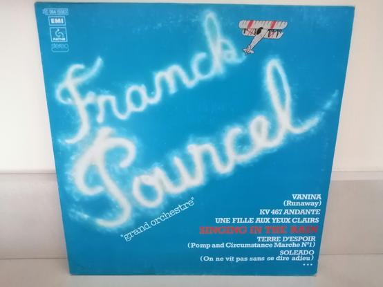 Sur un fond bleu, un avion biplan dessine le nom "Franck Pourcel" avec de la fumée.