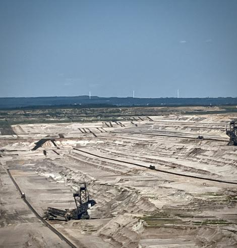 A wide panoramic view of a vast open-cast lignite mine, showing multiple layers of excavation, several large excavators, and a conveyor belt system in the foreground, bordered by green vegetation and a clear summer sky.