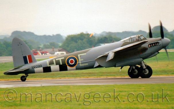 Side view of a twin propellor-engined, World War 2-era bomber aircraft taxiing along a runway from left to right.
The plane is covered in an olive-green and grey camouflage scheme, with thick black and white bands around the lower rear fuselage.
There are very pale letters "HT-E" either side of a blue, white, and red roundel outlined in yellow.
There is a blue, white, and red fin flash on the lower part of the tail, above the black serial "RR299" on the upper rear fuselage.
Lush green grass fills the foreground, with more grass leading up to a house and a fire truck in the distance, with trees beyond, under hazy grey skies.