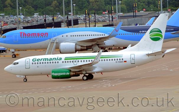 The white tail has a dark green circle with a lighter green slice in the middle.
The black registration "D-AGES" is on the lower rear fuselage next to a small German flag.
The engine pods slung under the wings are bright green, with white "FlyGermania.de" text.
The white upturned wingtips have that same text, but this time in green.
Grey concrete apron fills the foreground, with a larger, blue and white twin engined airliner stretching across the frame in the background, with a sprawling ground-level car park in front of trees in the distance.