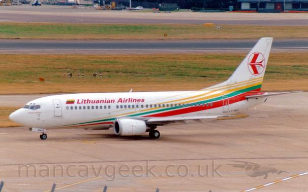 Side view of a twin engined jet airliner taxiing from right to left.
The plane is mostly white, with yellow, green, and red diagonal stripes running from the lower forward fuselage to the upper rear fuselage  and up into the lower reaches of the tail.
There are red "Lithuanian Airlines" on the upper forward fuselage, next to a small Lithuanian flag.
The black registration "LY-BAG" is on the lower rear fuselage.
The white tail has the red outline of a circle containing a stylised red flying bird.
Grey concrete apron fills the foreground, with wide swathes of grass either side of a black runway in the background.