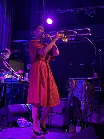 Laetitia Sadier of Stereolab in a red dress on stage, playing the trombone with another band member in the background.