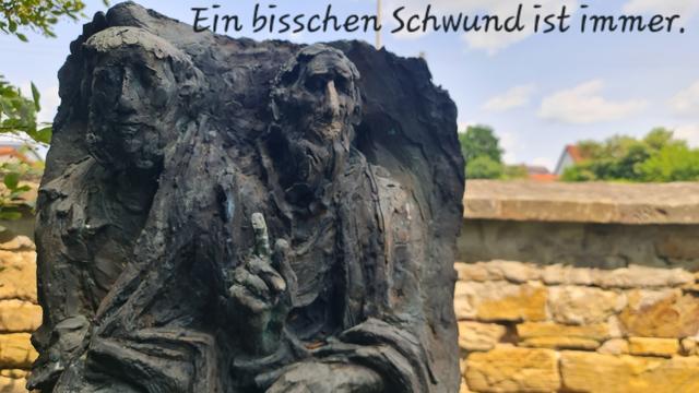 Relief-skulptur aus bronze vor einer steinmauer, zwei ältere Männer, vielleicht Pilger, der eine mit erhobenem Zeigefinger, spruch:
Ein bisschen Schwund ist immer.