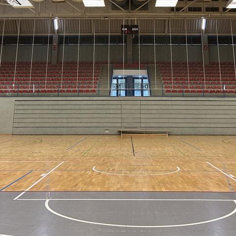 Eine leere Sporthalle, die Hardtberghalle in Bonn, mit Holzboden und markierten Linien für verschiedene Ballsportarten. Im Hintergrund befindet sich eine große Tribüne mit roten Sitzen, über der eine digitale Anzeigetafel hängt. Auf dieser steht »Heim 21 – Gäste 65«. Eine einzelne Holzbank steht an der Rückwand der Halle.