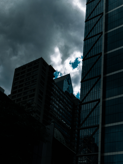 a dark shot of several buildings in a city. Bright blue sky peeks out from behind the clouds and buildings