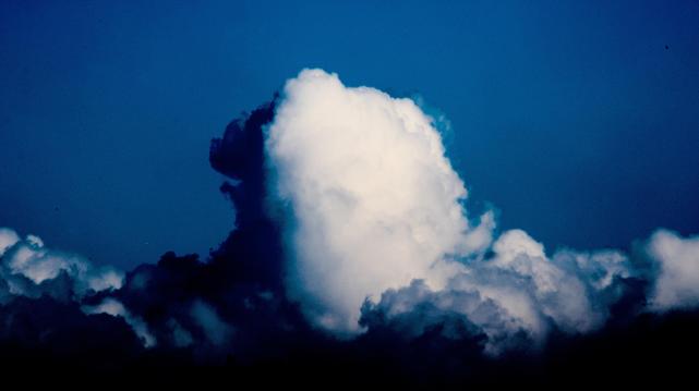 a heavily edited photo of a tall cumulus cloud. the cloud has a shadow on one side. the cumulus is sat in a bed of other clouds that get inky black near the bottom of the photo
