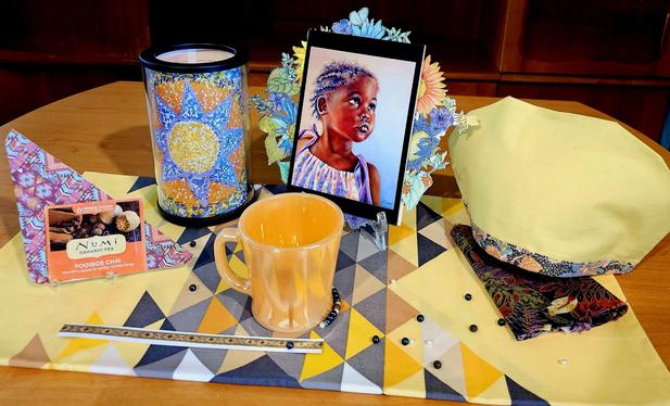 Tea tableau for Juneteenth centering on a portrait of a beautiful African-American child looking off into the distance, backed by a halo of wildflowers. There are African inspired textiles, a blazing sun mosaic candle, Rooibos Tea native to Africa, a gleaming orange FireKing mug, and loose beads and pearls scattered around like the Diaspora, with a separate section of beads held together by an invisible, unbroken cord.