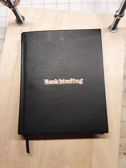 A black leather book with "Bookbinding " stamped on the cover with copper coloured foil. It lays on a book press on a work bench.