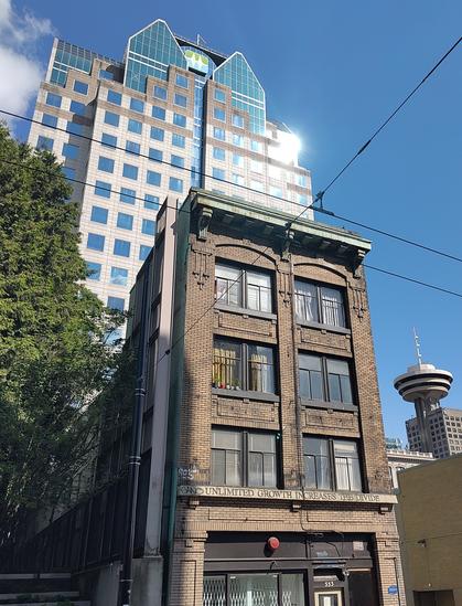 An old 4 storey building in brown brick fills the foreground. Behind, looms an enormous modern multistorey office building. The old building has the wording: 'Unlimited growth increases the divide' in weathered copper lettering.