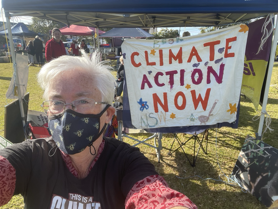 Masked woman wearing Climate Emergency tshirt standing in front of Climate Action Now sign