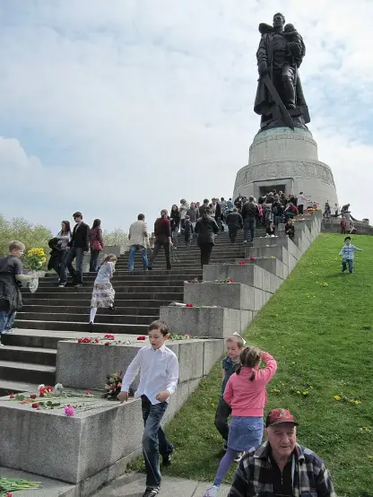 The image depicts a monument with a large sculpture at the top, surrounded by people walking up a set of stairs. There are flowers laid on the steps, and it appears to be an outdoor setting, possibly during a commemorative or memorial event. Children and adults are present, indicating a gathering or celebration related to the monument. The atmosphere seems lively, with many attendees participating in the event.

Image Credits: Wikimedia / Bernd.Brincken / CC BY-SA 3.0