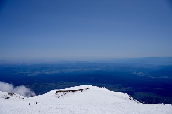 View from the summit looking south. The snow covered terrain drops quickly below to a saddle between the summit and Pikers Peak. The terrain is completely covered in snow except for some rocky ridges that stick up. The sky is clear and light blue. Some clouds can be seen below to the left of frame. The terrain around the mountain is distant and dark.