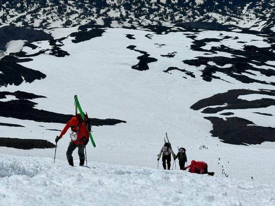View from the top of Pikers Peak. A number of people can be seen climbing up the steep snow covered face carrying packs and gear including skis. Lunch Counter is visible below with islands of dark volcanic rock sticking up through the vast snowfield. In the distance, evergreen trees can be seen sticking up from the snow at lower elevation.