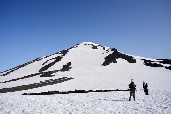 View looking up at Pikers Peak from Lunch Counter. A couple of people are visible in the foreground. A large number of people are visible climbing up in the background. The sky is light blue and clear. The terrain is mostly covered with snow except for large patches of volcanic rock.