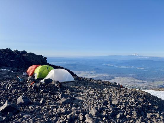 Camp at Lunch Counter. Three tents are pitched close together in a clearing of a rocky area that extends from left of frame. The tents are orange, green, and white. The terrain drops towards the right of frame with Mount Hood visible far off in the distance. The sky is light blue and clear.