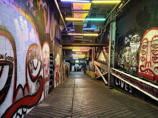 A view into a long hallway full of graffiti and painting with neon lights. One light says “XXX”, and at the end is “Jonathan Schwartz Productions” with some trash cans underneath.