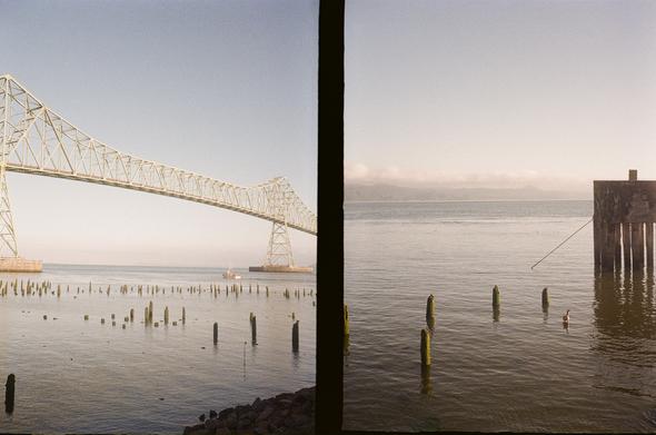 This is two photos on one frame of 35 mm film. The photo on the left is of a tall bridge arcing over a very wide river with a fishing boat going under it in the distance. The one on the right is half a curious concrete structure and some pilings. In both of the photos the colors are very muted and dreamy , as if it is early in the morning. The light is hazy and clouds are visible over the hills across the river. These were taken in Astoria Oregon about a month ago with a 1960s Russian camera called a Chaika II and some 200 ISO Kodak color film.