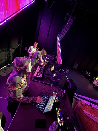 Seitenansicht auf die Bühne mit 3 Leuten. Ein VJ am Laptop, ein Mensch an midi Controllern und ein Saxophonist.