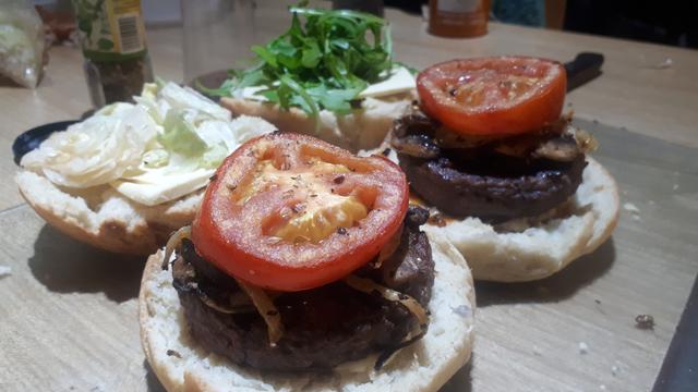 Two of the rolls, cut in half, laying on a cutting board on the kitchen table. grilled vegan burgers, cooked onions, mushrooms and tomatoes, lay on the 2 bottom baps, sliced cheese and garnishes lay on the top baps