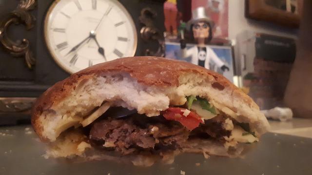 The filling can be seen inside of the second half eaten burger. A small statue of Alice Cooper can be seen peaking over from the background.
