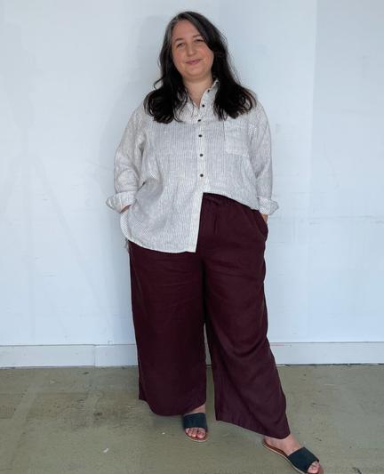 Caroline wears a relaxed pinstripe linen button-up, brown linen wide leg pants, and slides.