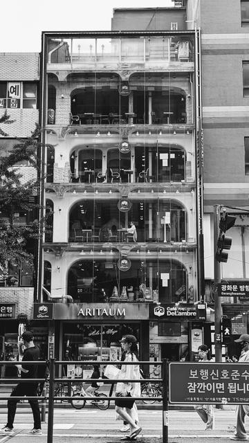 Black and white photo of a 5 level coffee shop in Seoul, South Korea. Image shows a lone customer on level 3 on his laptop. The windows are wide and open around a frame. Outside, locals go about their daily business.