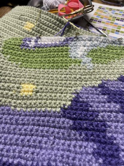 A close up of a crochet project featuring swaths of green moss and purple cliffs with the beginnings of a unicorn foot. This one has a darker purple and lighter blue.
