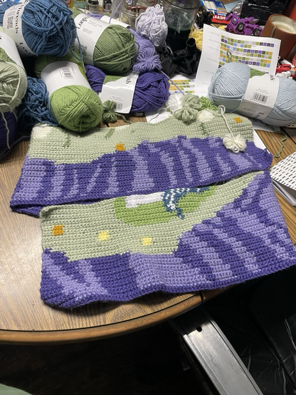 A close up of a crochet project featuring swaths of green moss and purple cliffs with the beginnings of a unicorn foot. This one has a lighter purple and darker blue.