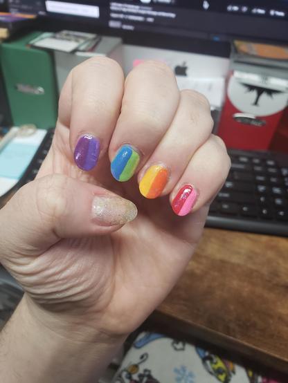 a photo of my painted nails, the four fingers each have two colours (split vertically down the middle), using all 8 colours of Gilbert Baker's pride flag. the thumb is a mix of holographic and opalescent sparklies