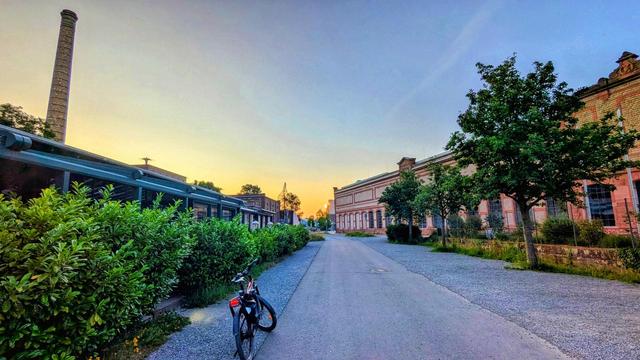 Eine ruhige Straße bei Sonnenaufgang, gesäumt von alten Industriegebäuden aus Backstein und Grünanlagen. Links steht ein Fahrrad an einer Hecke, rechts stehen Bäume vor einem historischen Fabrikgebäude. Im Hintergrund ist ein hoher Schornstein zu sehen.