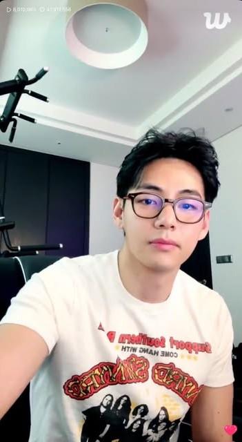 V of BTS seated in his gym/game room. He is wearing a rock band shirt and glasses.