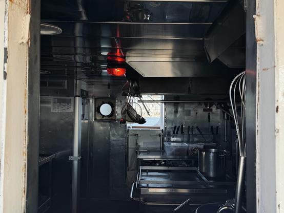 An interior view of a ship’s kitchen (galley), with stainless steel surfaces, cooking equipment, and knives visible on the wall. A red light illuminates the space, and there's a circular window showing an outdoor view.