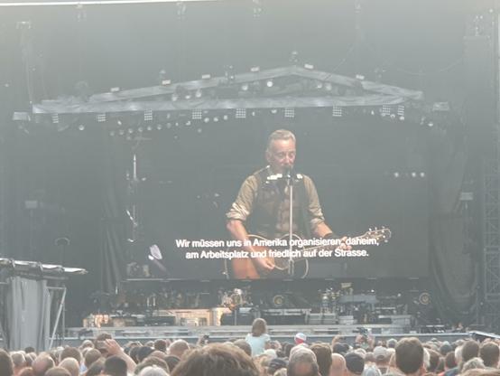 Foto. Bruce Springsteen im Frankfurter Waldstadion auf der Bühne. Er hat eine Akustikgitarre umhängen und spricht ins Mikrofon. Weil ich ziemlich weit hinten stand, ist der Boss nur sehr winzig zu sehen, über der Bühne ist aber ein riesiger Bildschirm, auf dem er auch in groß zu sehen ist. Vor der Bühne jede Menge Leute. Zu Springsteens politischen Zwischenansprachen sind auf dem Bildschirm deutschsprachige Untertitel eingeblendet. Hier: "Wir müssen uns in Amerika organisieren, daheim, am Arbeitsplatz und friedlich auf der Straße".