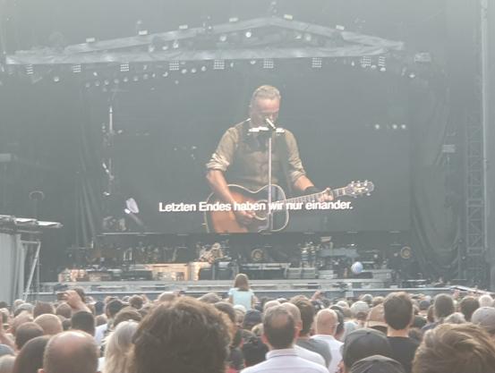 Foto. Bruce Springsteen im Frankfurter Waldstadion auf der Bühne. Er hat eine Akustikgitarre umhängen und spricht ins Mikrofon. Weil ich ziemlich weit hinten stand, ist der Boss nur sehr winzig zu sehen, über der Bühne ist aber ein riesiger Bildschirm, auf dem er auch in groß zu sehen ist. Vor der Bühne jede Menge Leute. Zu Springsteens politischen Zwischenansprachen sind auf dem Bildschirm deutschsprachige Untertitel eingeblendet. Hier: "Letzten Endes haben wir nur einander".