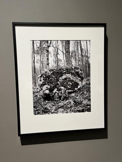 Photo of a framed black and white photograph. In it, a black man in a military uniform and bare feet sits hunched over in front of a large boulder. The boulder has lots of contrast, perhaps lichen or moss or paint, which evokes his camouflage. Trees rise up behind the boulder.