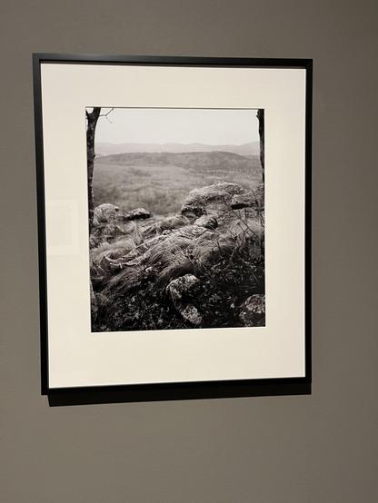In this black and white Framed photograph, there is a view of nature from a hill. In the distance is a hilly landscape, up close a rocky and grassy hillside. It took me a moment to find him in this photo, as his camouflage uniform hides him very well with the grass. His head and his feet could be rocks at first glance. But once you see him, lying flat, at a rather steep angle, it becomes visceral.