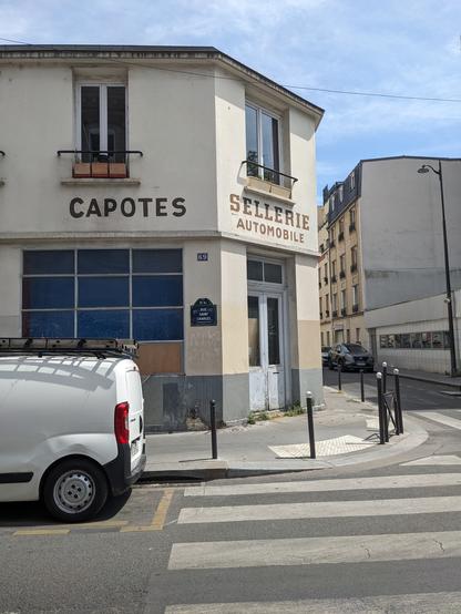 Au coin d'une rue parisienne, un immeuble d'un étage siège d'un atelier de Sellerie automobile (en lettres peintes brunes, sur le pignon). Également en lettres peintes, la mention Capotes. Deux plaques émaillées : 69 et rue Saint-Charles.